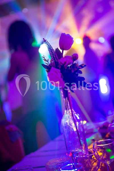 Un vase en verre contenant des fleurs blanches et roses sur une table, avec des lumières colorées en arrière-plan.