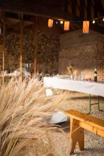 Décoration rustique avec épis de blé au premier plan, tables dressées et guirlandes lumineuses dans une grange.