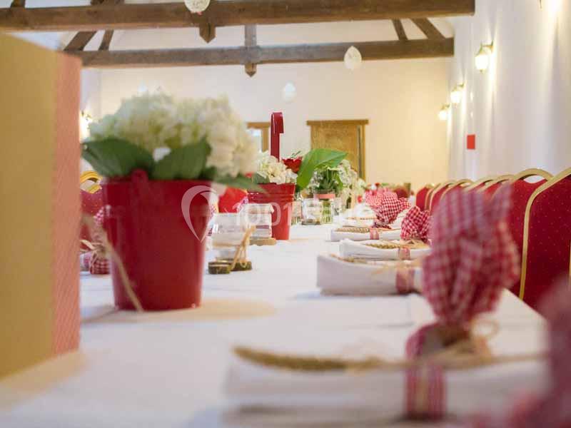 Table décorée avec des nappes blanches, pots de fleurs rouges et serviettes pliées, dans une salle aux poutres apparentes.