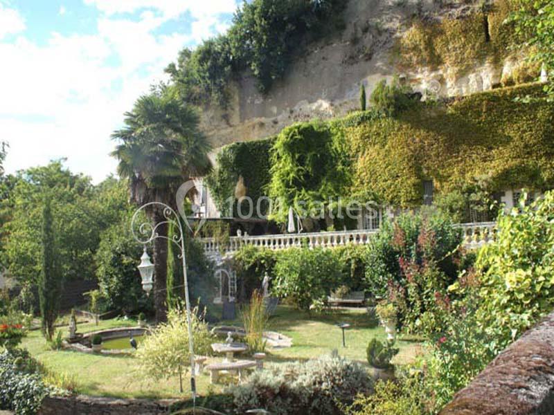 Jardin verdoyant avec terrasse ombragée, façade recouverte de végétation et falaise en arrière-plan.