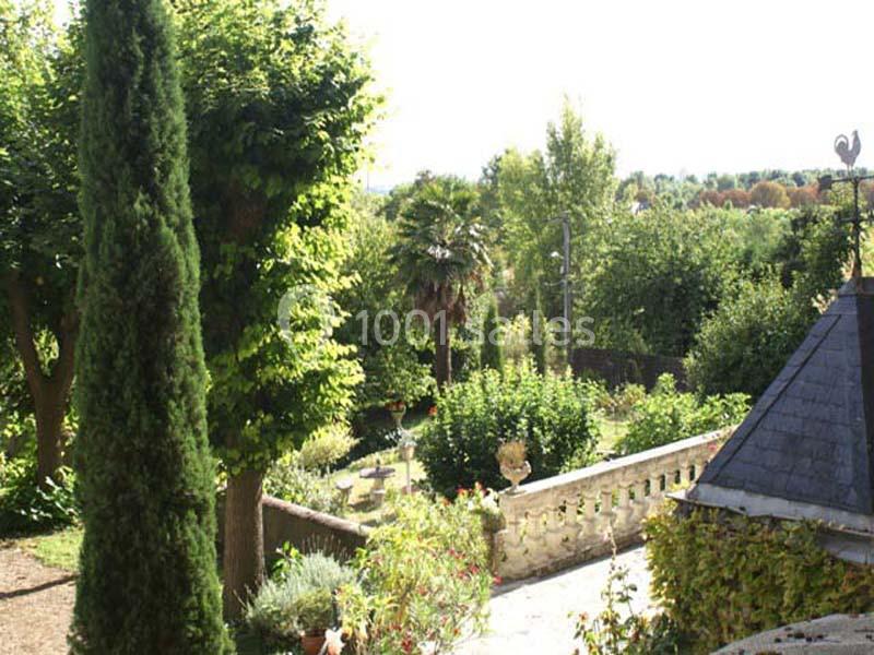 Jardin verdoyant avec arbres, arbustes et terrasse en pierre, sous un ciel clair et lumineux.