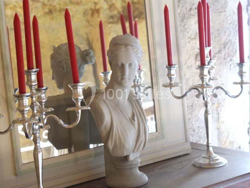 Buste en marbre blanc entouré de chandeliers argentés avec bougies rouges, posé devant un miroir.