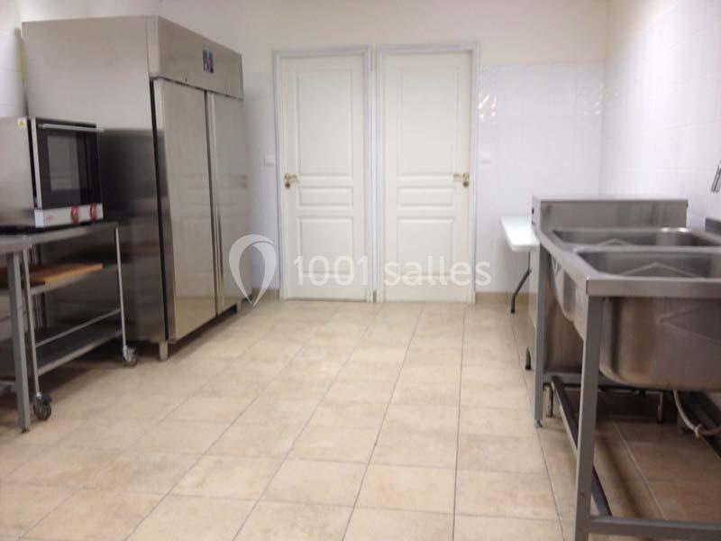 Cuisine professionnelle avec sol carrelé, évier inox, réfrigérateur, four et murs blancs.