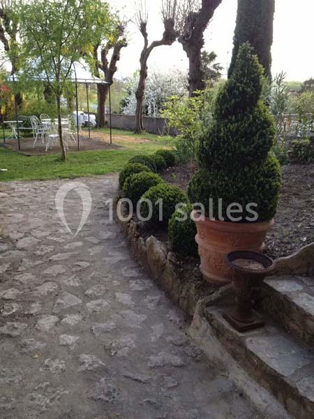 Allée en pierre bordée de buis taillés, avec un pot en terre cuite et un jardin arboré en arrière-plan.