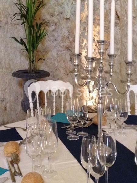 Table dressée avec nappes blanches et bleues, chandeliers argentés, verres et pains, dans un décor en pierre avec plante en…