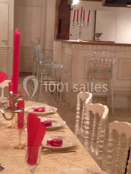 Table dressée avec des bougies rouges, serviettes pliées et assiettes, devant un bar avec des tabourets transparents.