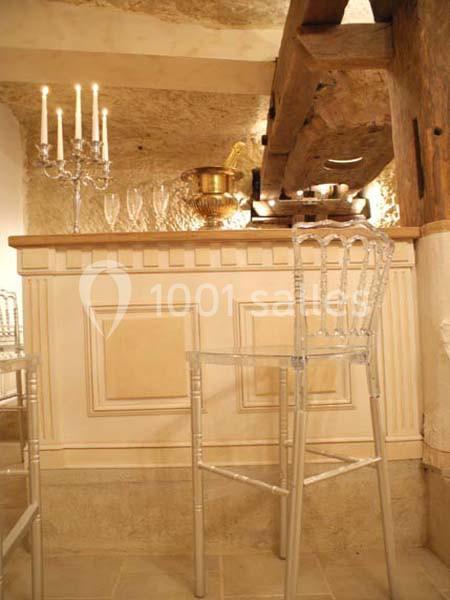 Bar en bois clair avec un comptoir décoré de chandeliers, verres et un seau à champagne, entouré de chaises transparentes.