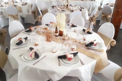 Table ronde décorée pour un événement, avec nappes beige, chaises blanches ornées de tissu orange et centre de table floral.