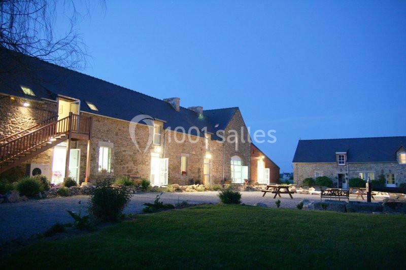 Bâtiments en pierre éclairés de nuit, entourés de végétation et d'une cour avec des bancs en bois.