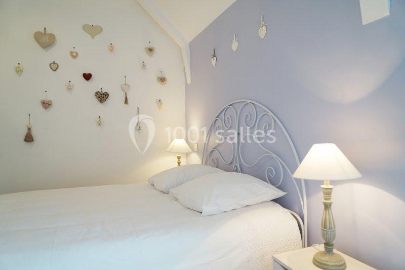 Lit avec tête de lit en métal, draps blancs, mur violet décoré de cœurs suspendus, éclairé par deux lampes de chevet.