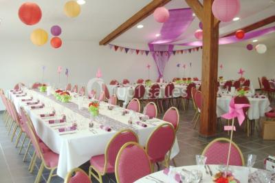 Table ronde décorée pour un événement, avec nappes beige, chaises blanches ornées de tissu orange et centre de table floral.