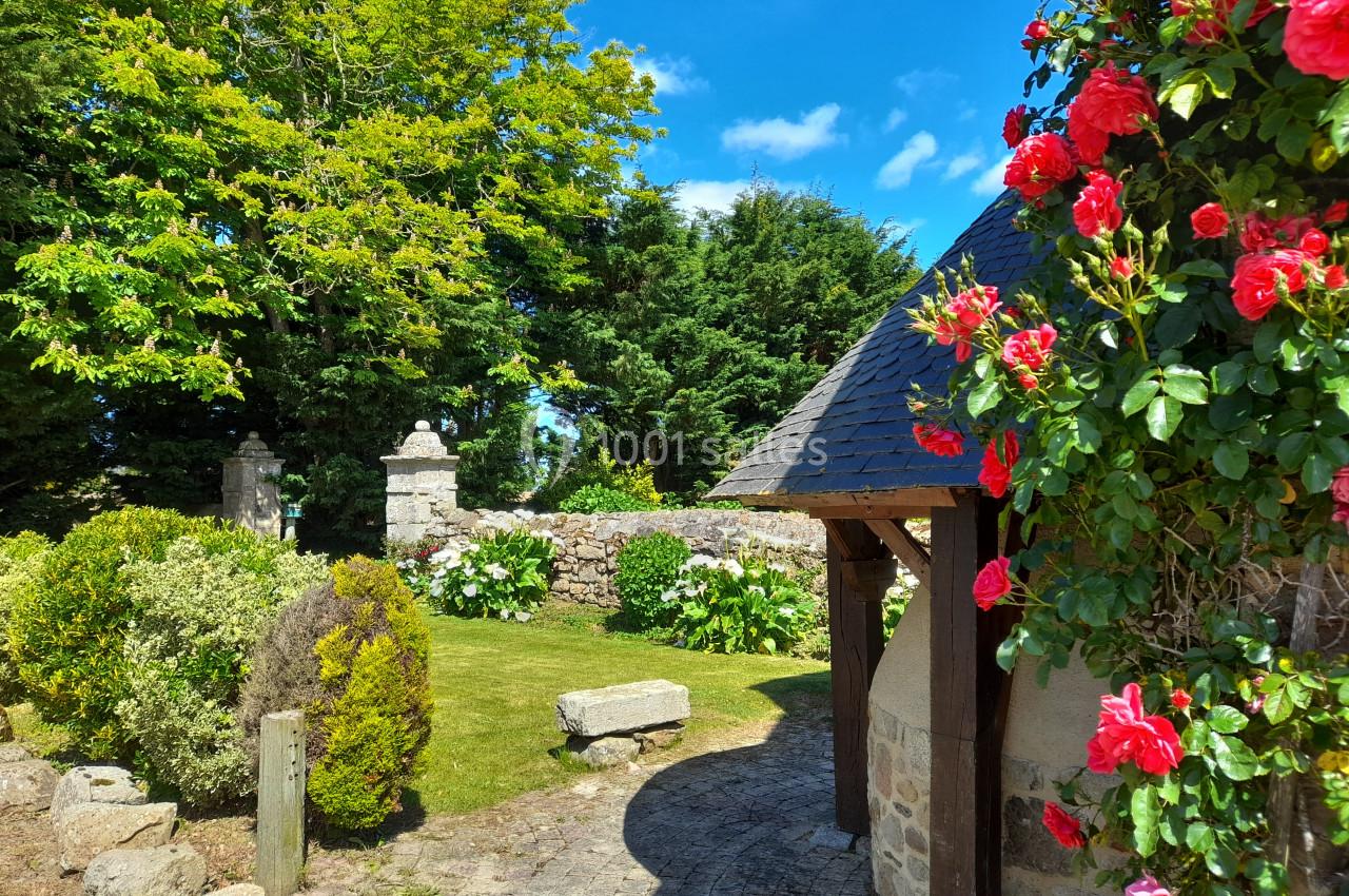 Jardin fleuri avec rosiers rouges, pelouse, muret en pierre et arbres sous un ciel bleu ensoleillé.
