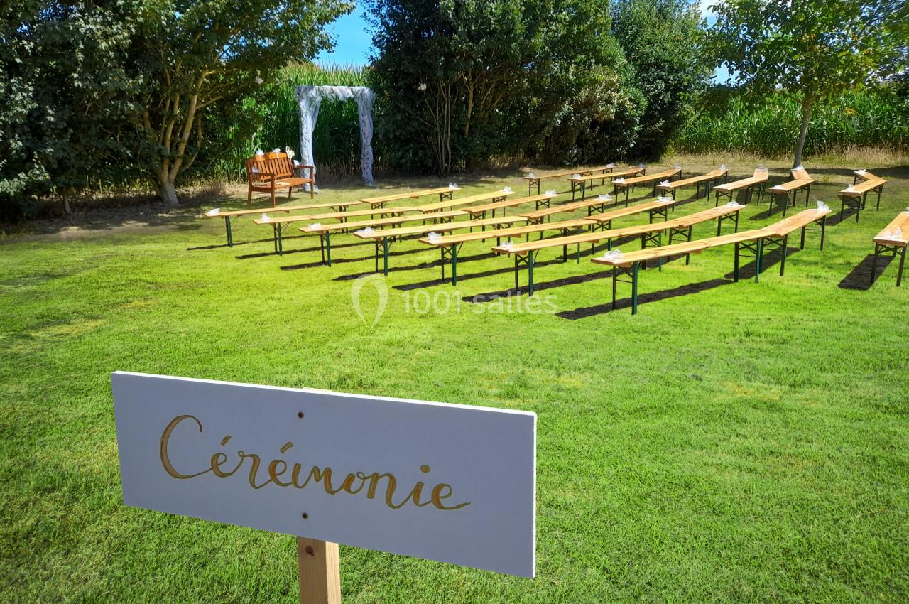 Bancs en bois alignés en extérieur pour une cérémonie, avec une arche décorative et un panneau ’Cérémonie’.