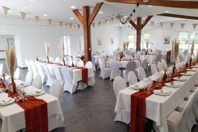 Table ronde décorée pour un événement, avec nappes beige, chaises blanches ornées de tissu orange et centre de table floral.