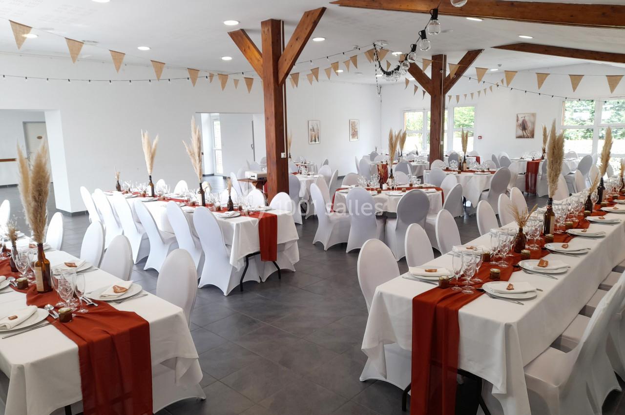 Salle de réception décorée pour un événement, avec tables dressées, nappes blanches et chemins de table rouges.