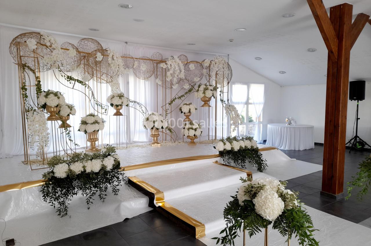 Décoration de scène de mariage avec fleurs blanches, arches dorées et rideaux blancs dans une salle lumineuse.