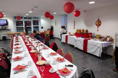 Table ronde décorée pour un événement, avec nappes beige, chaises blanches ornées de tissu orange et centre de table floral.
