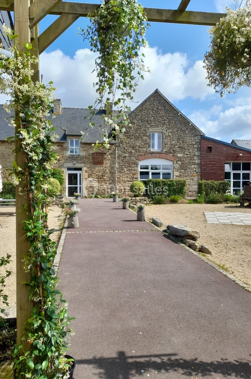 Allée pavée bordée de plantes menant à une bâtisse en pierre sous un ciel partiellement nuageux.