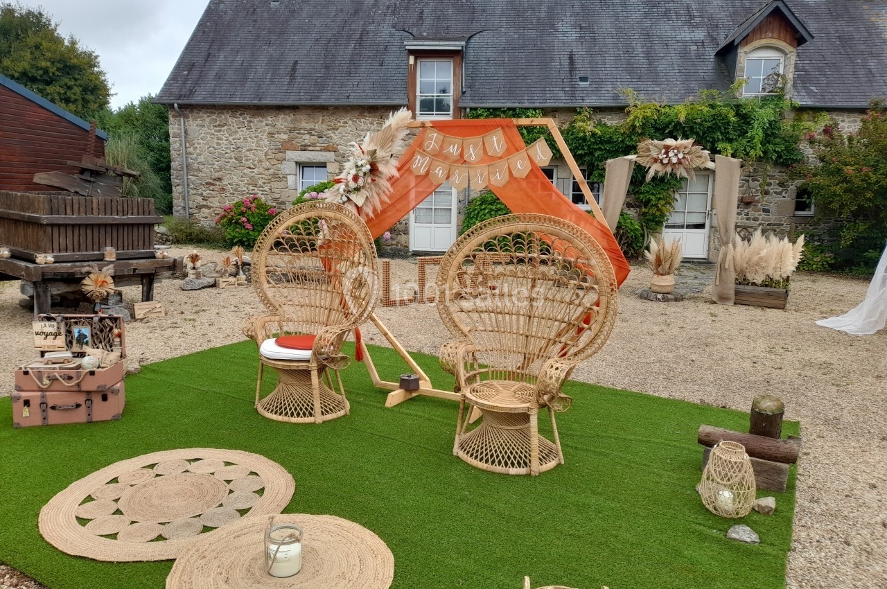 Espace extérieur décoré pour un événement avec fauteuils en rotin, arche orange et éléments bohèmes devant une maison en…