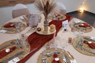 Table ronde décorée pour un événement, avec nappes beige, chaises blanches ornées de tissu orange et centre de table floral.