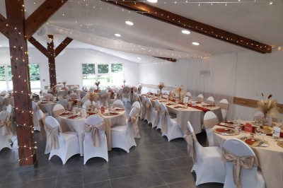 Table ronde décorée pour un événement, avec nappes beige, chaises blanches ornées de tissu orange et centre de table floral.