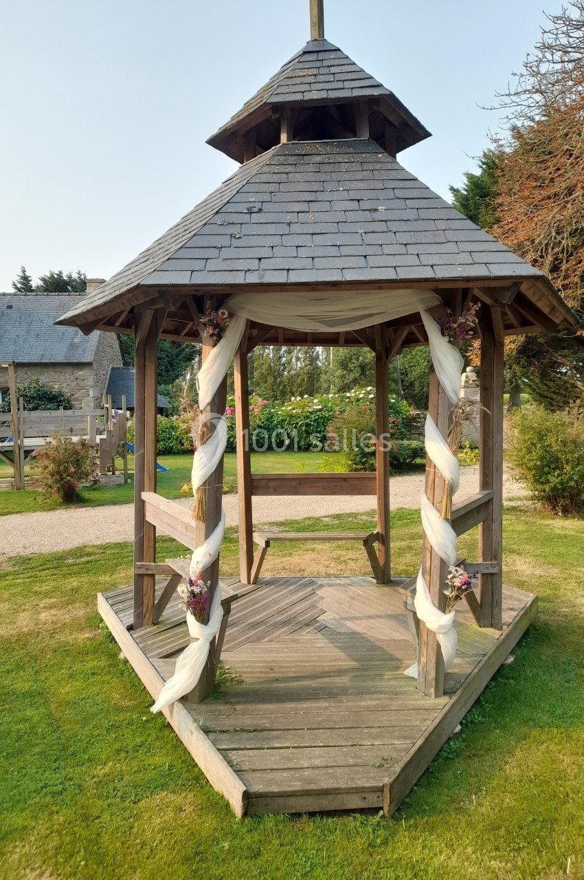 Kiosque en bois décoré de voiles blancs et de fleurs, situé sur une pelouse dans un jardin.