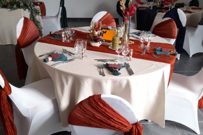 Table ronde décorée pour un événement, avec nappes beige, chaises blanches ornées de tissu orange et centre de table floral.