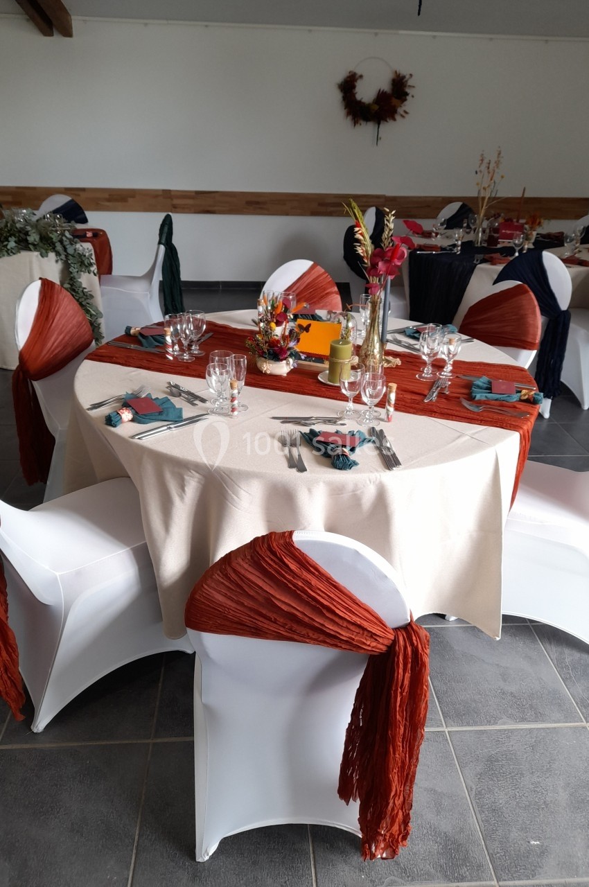Table ronde décorée pour un événement, avec nappes beige, chaises blanches ornées de tissu orange et centre de table floral.