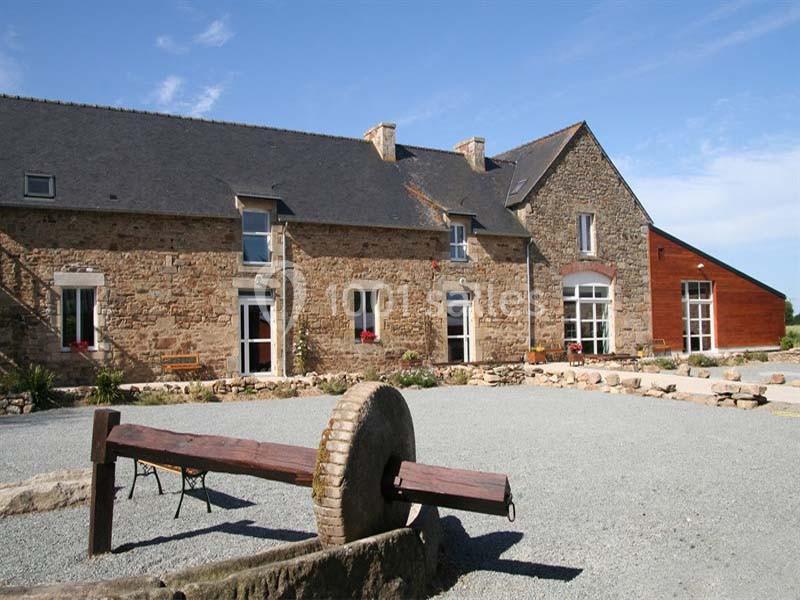 Bâtisse en pierre avec toit en ardoise, fenêtres modernes et une ancienne meule en pierre au premier plan.