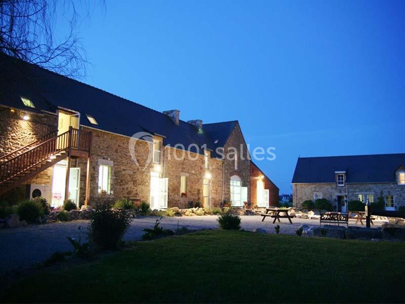 Bâtiments en pierre éclairés la nuit, entourés de végétation et d'une cour avec des tables de pique-nique.