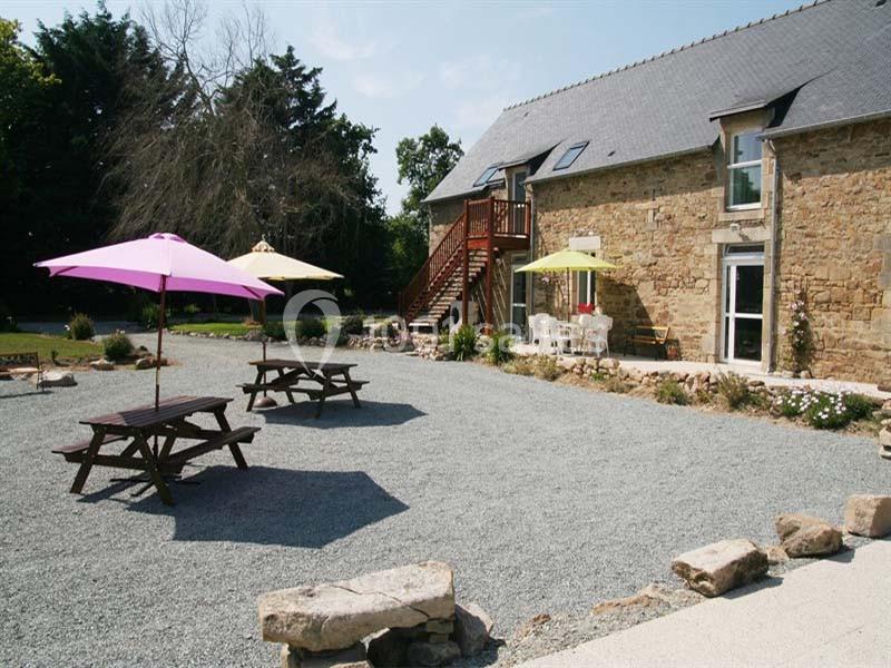 Cour en gravier avec tables de pique-nique, parasols colorés et bâtiment en pierre avec escalier en bois.
