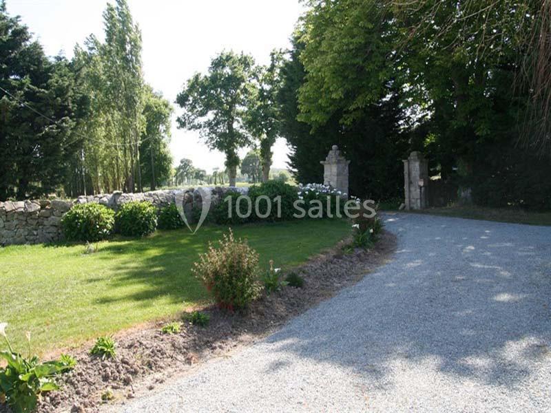 Allée gravillonnée bordée de pelouse, arbustes et arbres, menant à une entrée avec piliers en pierre.