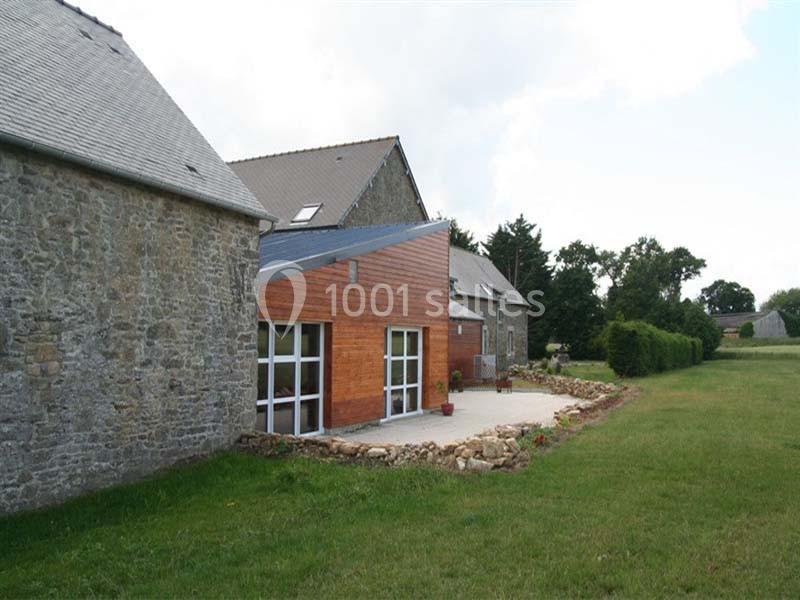 Façade d'une maison en pierre et bois avec terrasse, entourée de pelouse et d'arbres en arrière-plan.