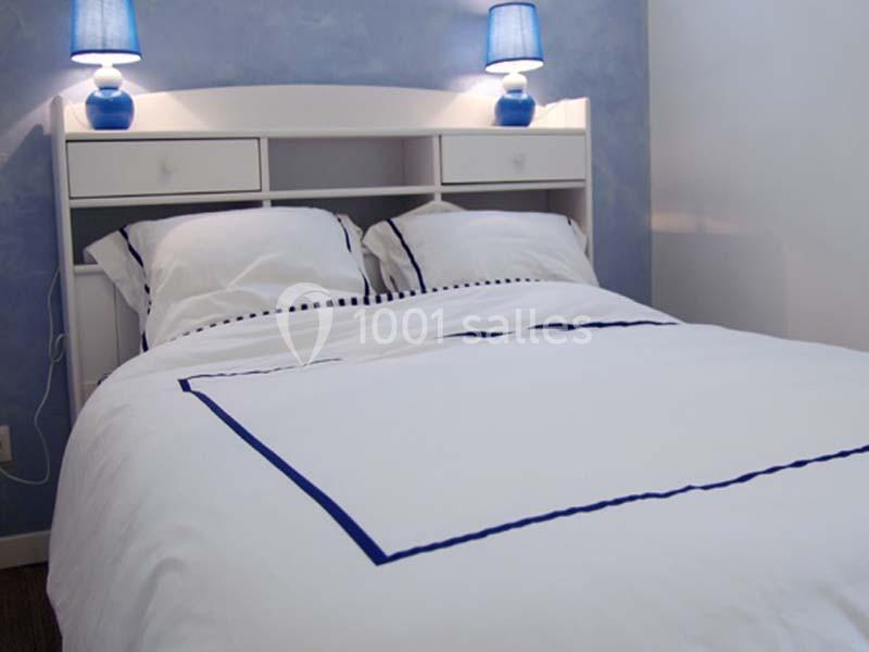 Lit double avec draps blancs bordés de bleu, encadré par deux lampes de chevet sur une tête de lit blanche.