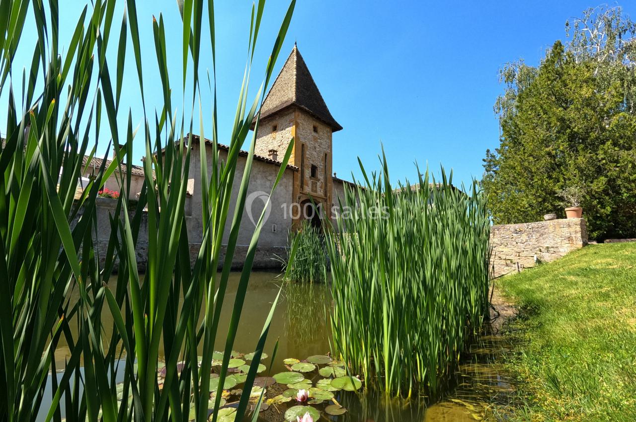 Vue d'un bâtiment ancien avec une tour, entouré de roseaux et d'un étang avec des nénuphars sous un ciel bleu.