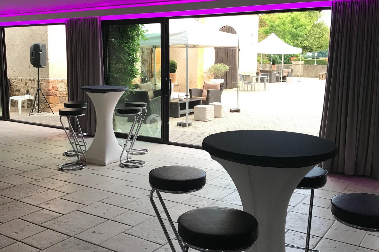 Salle avec tables hautes et tabourets, donnant sur une terrasse extérieure aménagée visible à travers de grandes baies…