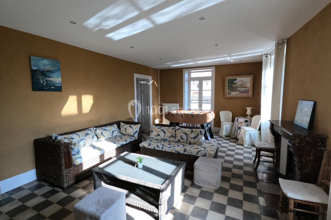 Salon lumineux avec canapé en rotin, table basse en verre, murs ocres, cheminée et sol en carreaux noirs et blancs.