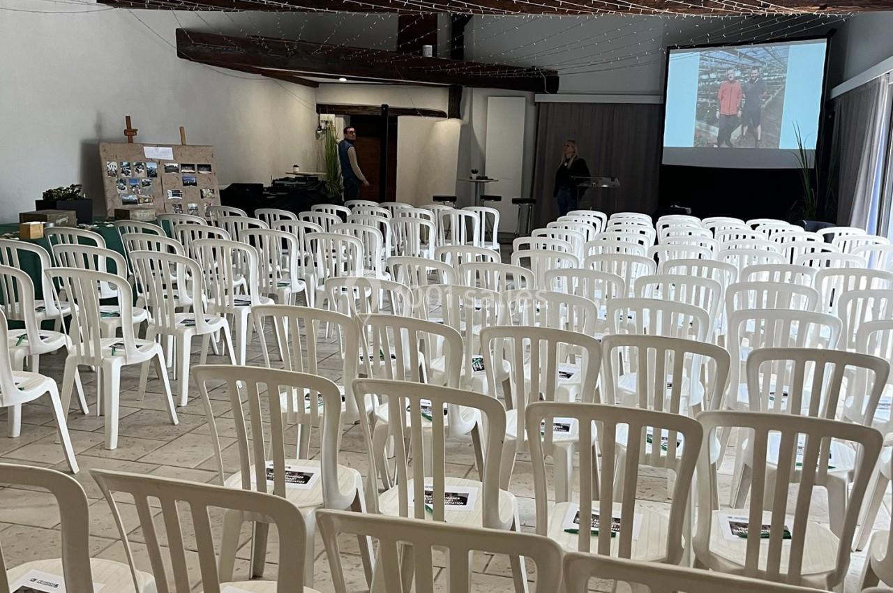 Salle de conférence avec des rangées de chaises blanches vides, un écran de projection et des présentateurs en arrière-plan.