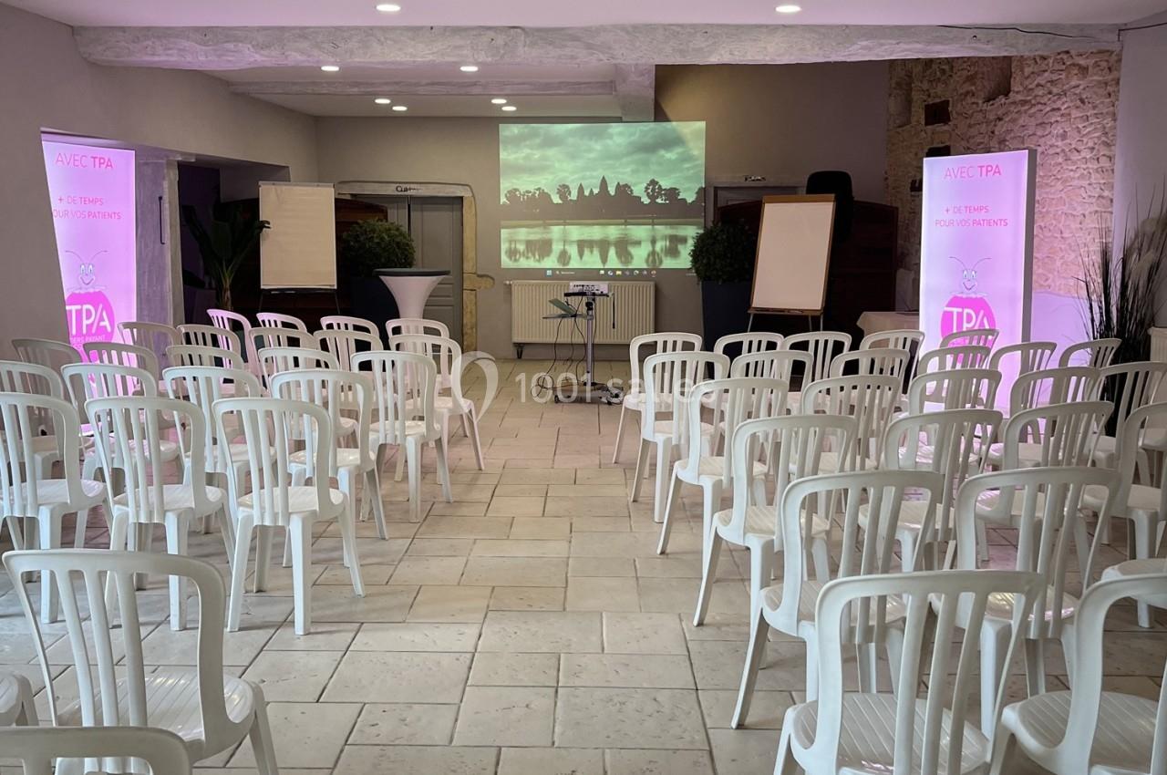 Salle de conférence avec des rangées de chaises blanches, un écran de projection allumé et des affiches promotionnelles.
