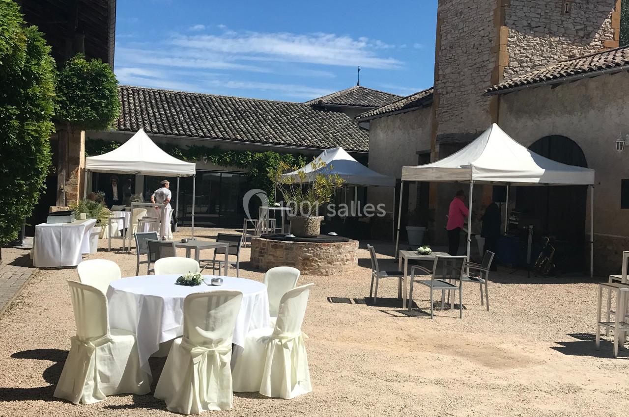 Cour extérieure ensoleillée avec tables dressées et chaises blanches, entourée de bâtiments en pierre et de tentes blanches.