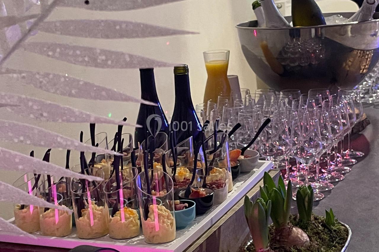Buffet avec verrines, bouteilles de vin, jus d’orange, verres et décoration végétale sur une table éclairée.