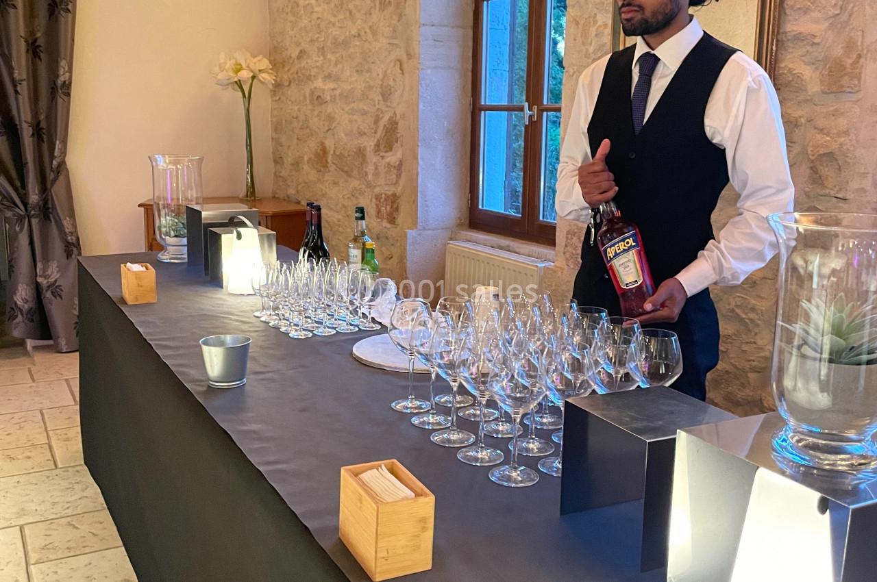Un serveur en tenue formelle prépare un bar avec des verres à vin alignés dans une salle en pierre éclairée.