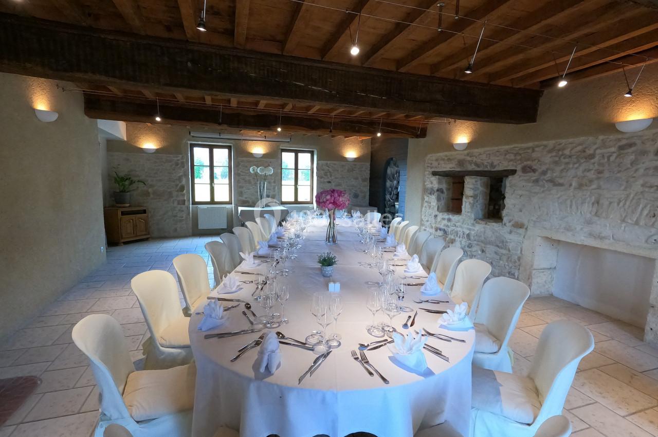 Salle de réception avec une grande table ovale dressée, entourée de chaises blanches, sous un plafond en bois.
