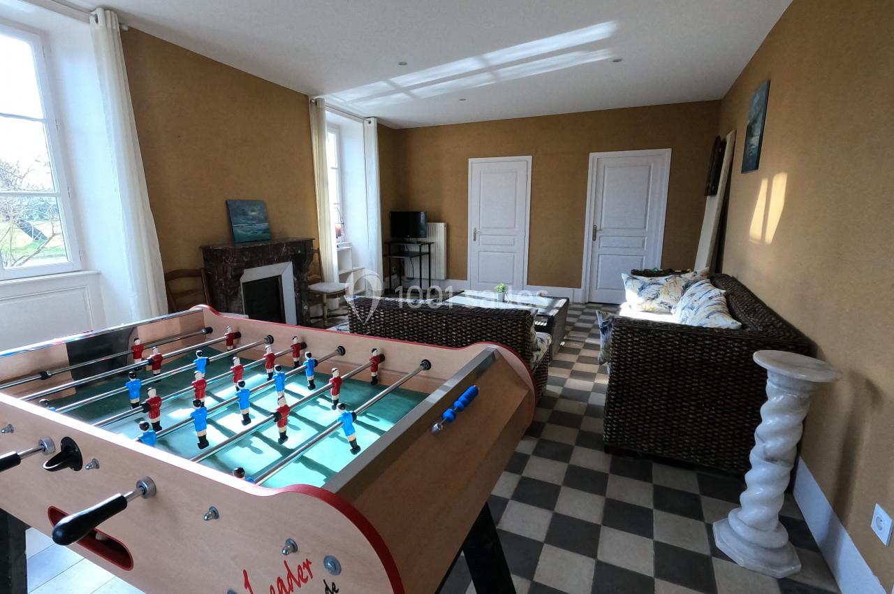 Salle de séjour avec baby-foot au premier plan, canapé, télévision et cheminée dans un intérieur lumineux.