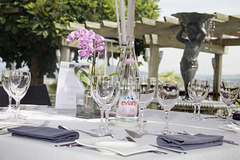 Table dressée avec des verres, une bouteille d'eau, des serviettes pliées et une orchidée, en extérieur près d'une statue.