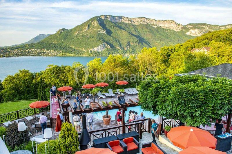 Terrasse avec piscine, parasols rouges, vue sur un lac entouré de montagnes et végétation luxuriante.