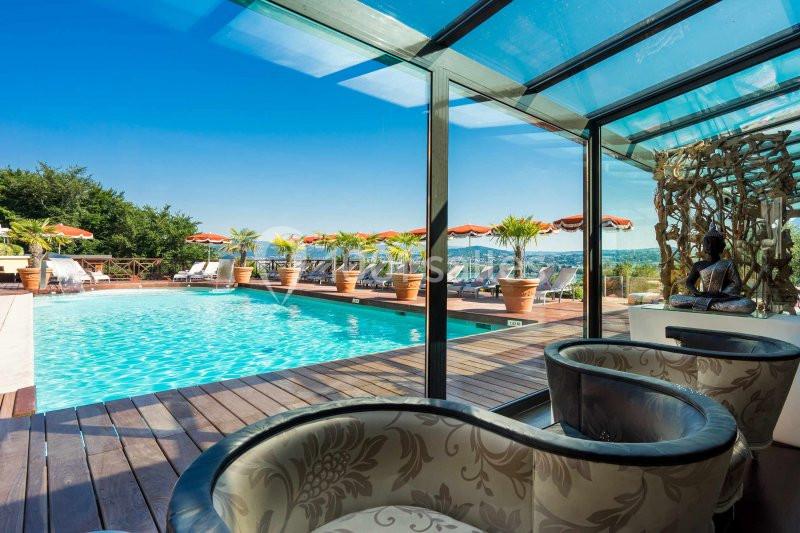 Vue sur une piscine extérieure entourée de transats et de parasols, avec un coin salon intérieur en premier plan.