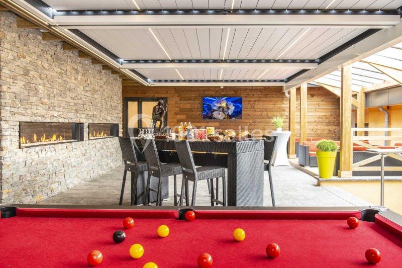 Salle avec billard rouge, table haute dressée, murs en pierre et bois, cheminée moderne et écran mural.