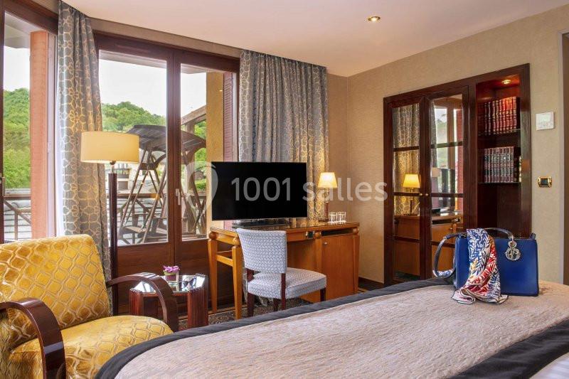 Chambre lumineuse avec lit, fauteuil, bureau, télévision et balcon donnant sur un paysage verdoyant.