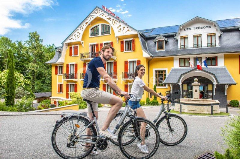 Un homme et une femme à vélo devant un hôtel jaune avec des volets rouges et une fontaine.